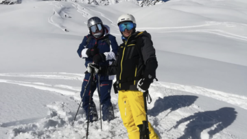 Zwei Skifahrer stehen im frischen Schnee unter klarem Himmel. Im Hintergrund sind schneebedeckte Berge zu sehen.