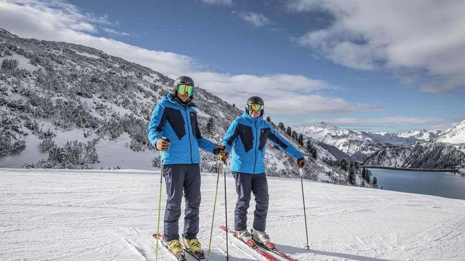Zwei Skifahrer stehen auf einer schneebedeckten Piste. Im Hintergrund sind Berge und ein klarer Himmel zu sehen.