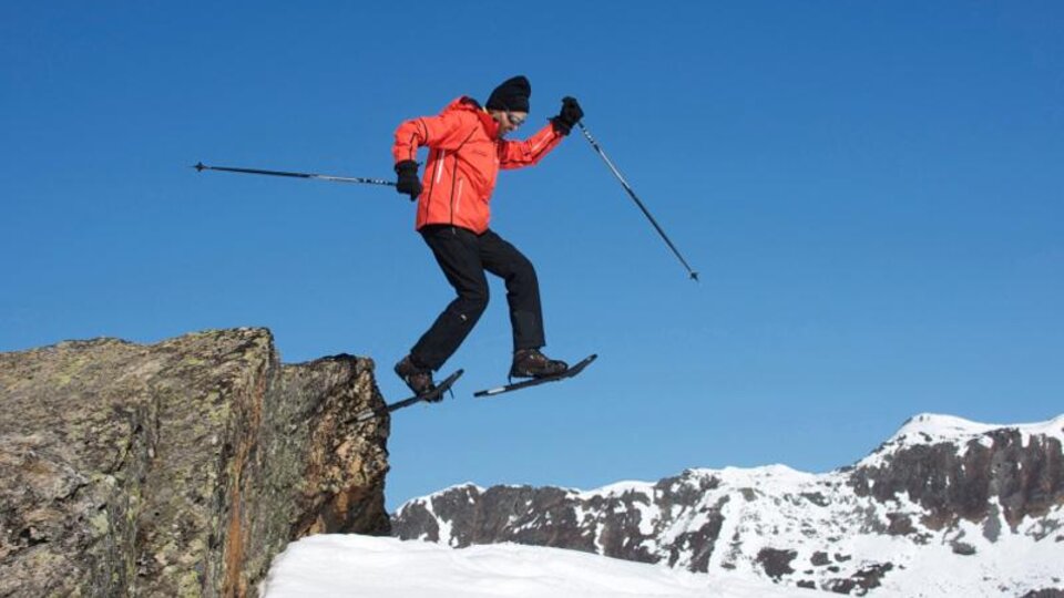 A man in a bright orange jacket jumps over a rock in the snow. In the background, snow-covered mountains and a clear blue sky can be seen.