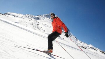 A person in red ski gear is skiing on a snow-covered slope. In the background, snow-covered mountains and a clear blue sky can be seen.