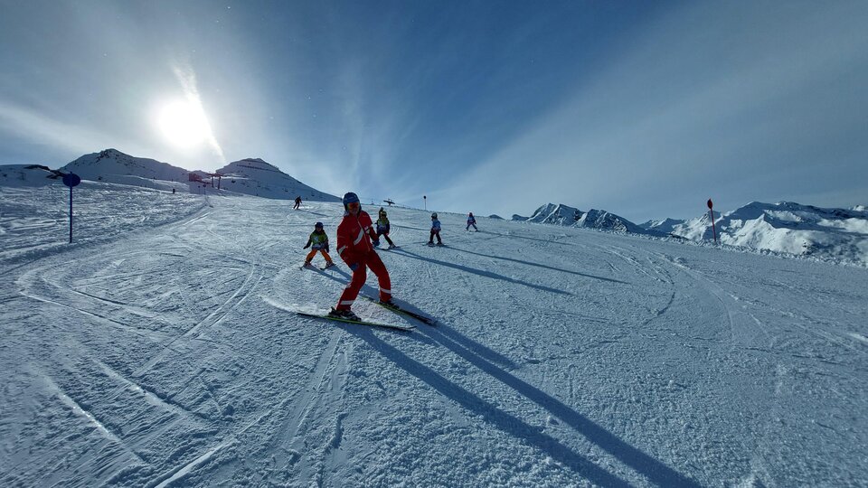 Eine Gruppe von skifahrenden Personen auf einer sonnigen Piste. Im Hintergrund sind schneebedeckte Berge zu sehen.