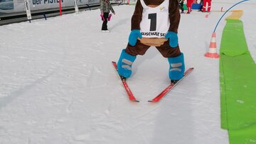 Ein Ski-Maskottchen steht auf Skiern im Schnee. Im Hintergrund sind Kinder und Skilehrer zu sehen.
