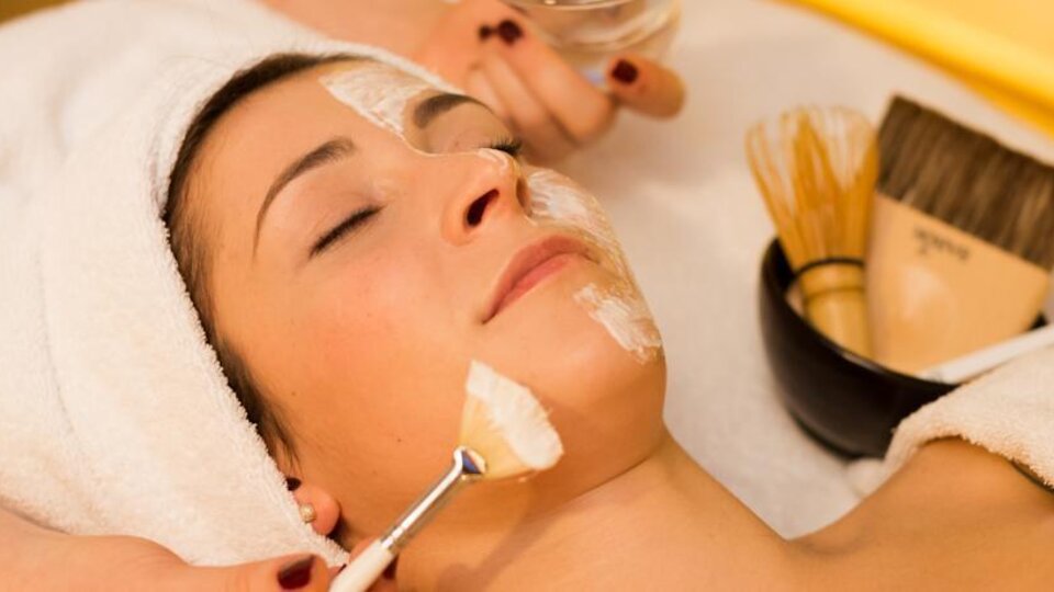 Eine Frau erhält eine Gesichtsbehandlung mit einer Maske auf ihrem Gesicht. Um sie herum sind verschiedene Beauty-Werkzeuge und eine Schüssel sichtbar.