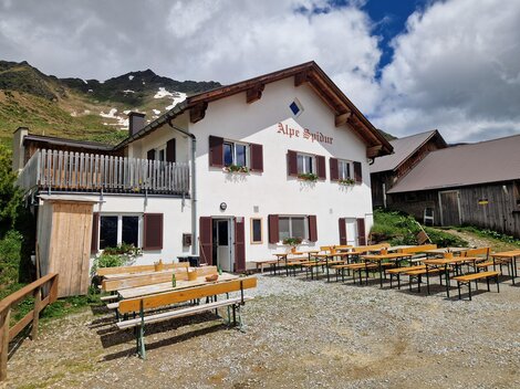 Ein gemütliches Alpenhaus mit dem Namen "Alpe Spina". Draußen stehen viele Bänke und Tische in einer malerischen Berglandschaft.