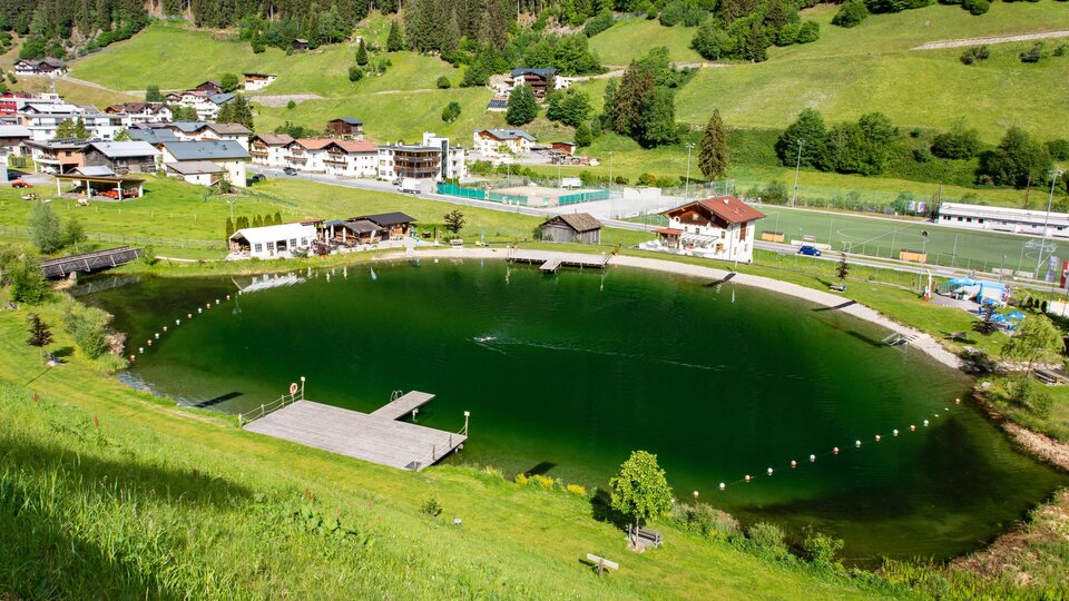 Ein malerischer Teich umgeben von grünen Wiesen und Bergen. Im Hintergrund sind mehrere Häuser und Anlagen sichtbar.