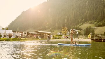 Eine Person steht auf einem Stand-Up-Paddle-Board auf einem ruhigen See. Im Hintergrund sind Berge und ein gemütliches Gebäude zu sehen, während die Sonne scheint.