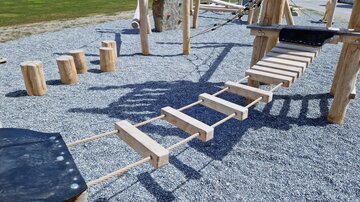 Eine Spielplatzbrücke aus Holz mit Seilen, die über eine Kiesfläche führt. Im Hintergrund sind weitere Spielgeräte und eine grüne Fläche sichtbar.