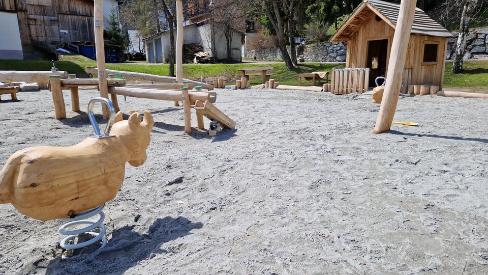 Ein Spielplatz mit einem Holzspringpferd und einem Spielhaus. Der Boden besteht aus Sand und es gibt weitere Spielgeräte in der Nähe.