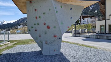 Eine moderne Kletterwand im Freien mit bunten Griffen. Sie steht auf einem Kiesplatz vor einer landschaftlichen Kulisse.