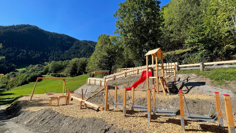 Ein Spielplatz in der Natur mit Holzelementen und einer Rutsche. Im Hintergrund sind grüne Hügel und Bäume zu sehen.