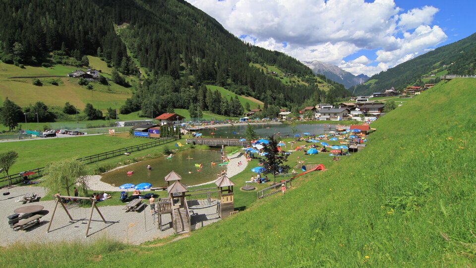 Ein malerischer Park mit einem Badesee und grünen Wiesen. Im Hintergrund sind Berge und Häuser zu sehen, während Menschen entspannen und die Sonne genießen.