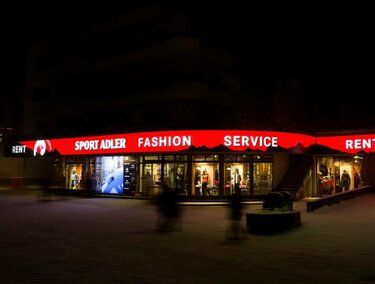 A shop with a bright red sign displaying "Sport Adler" and "Fashion Service". The scene is at night, and blurred figures can be seen in the foreground.