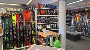Ein Geschäft für Wintersportartikel mit vielen Skiern und Skischuhen. Die Regale sind gut gefüllt und es gibt einen gemütlichen Sitzbereich.