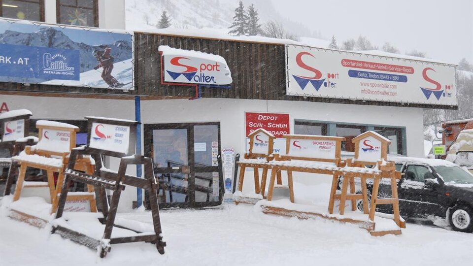 A sports store in winter with lots of snow in front of the door. The facade displays signs and wooden racks for ski equipment.