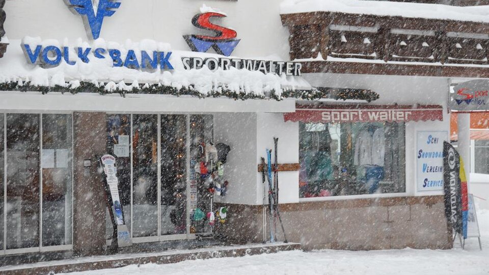 Ein Sportgeschäft mit Skiausrüstung und einer Bank im Winter. Dicke Schneeflocken fallen und die Umgebung ist verschneit.