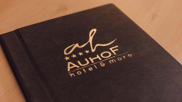 An elegant notebook with the logo of the hotel "Auhof". It has a dark surface and golden inscriptions.