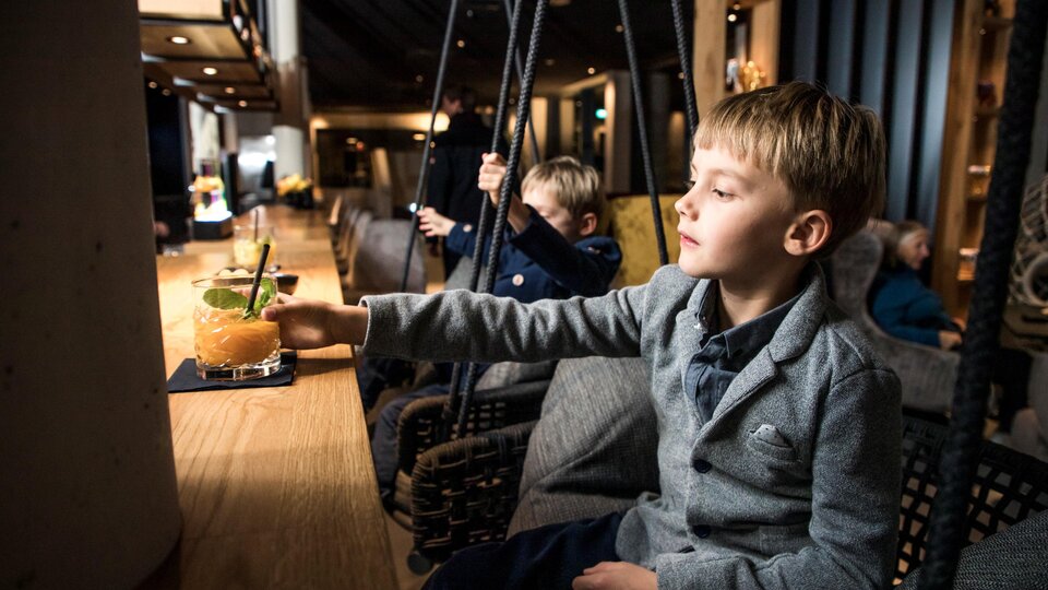 Ein Junge in einem schlichten grauen Anzug sitzt an der Bar und greift nach einem Getränk. Im Hintergrund sind weitere Kinder und eine gemütliche Innenausstattung sichtbar.