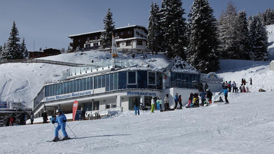 Ein schneesicherer Skikurs mit einem modernen Restaurant im Vordergrund. Viele Menschen genießen aktiv den Winterspaß in einer malerischen Berglandschaft.