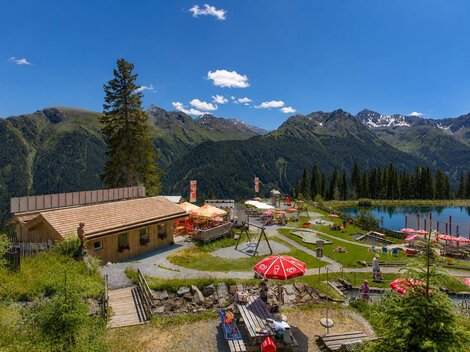 Eine alpine Freizeitanlage vor beeindruckender Bergkulisse: Im Vordergrund laden Holzterrasse, Liegewiese, Spielplatz und rote Sonnenschirme zum Verweilen ein, während rechts ein klarer Bergsee die umliegenden Gipfel spiegelt. Das helle Holzgebäude, die grünen Wiesen und der weite blaue Himmel vermitteln eine sommerliche, familienfreundliche Atmosphäre mitten in den Alpen.