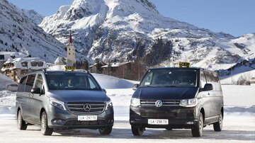 Zwei Taxis stehen im Schnee vor einer beeindruckenden Berglandschaft. Im Hintergrund sind schneebedeckte Berge und ein kleines Dorf zu sehen.