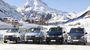 Eine Reihe von Taxis steht vor einer schneebedeckten Berglandschaft. Die Fahrzeuge sind in verschiedenen Farben und Modellen angeordnet.