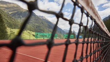 Ein Tennisplatz mit rotem Belag, umgeben von Bergen und Bäumen. Das Netz im Vordergrund gibt den Blick auf die Spieloberfläche frei.