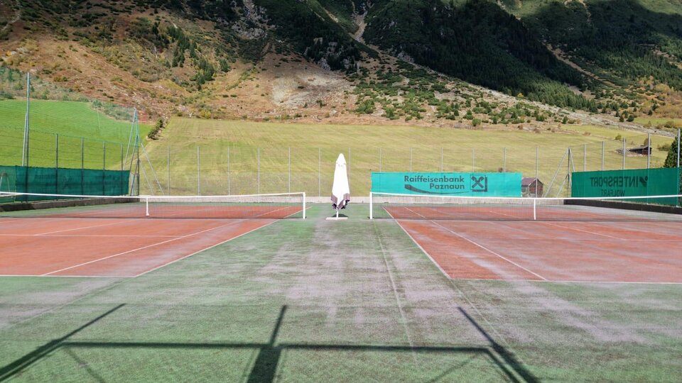 Ein Tennisplatz in einer malerischen Berglandschaft. Die Umgebung ist grün und sonnig mit klar blauen Himmel.