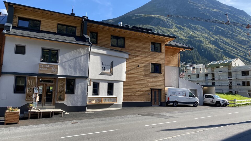 Ein modernes Gebäude mit Holzverkleidung und großen Fenstern in einer Berglandschaft. Im Hintergrund ist ein hoher Berg unter blauem Himmel sichtbar.