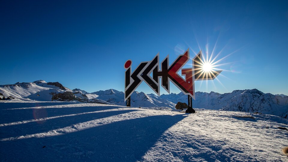 A beautiful viewpoint in Ischgl with a large sign and snow-covered mountains. The sun shines in the clear blue sky.