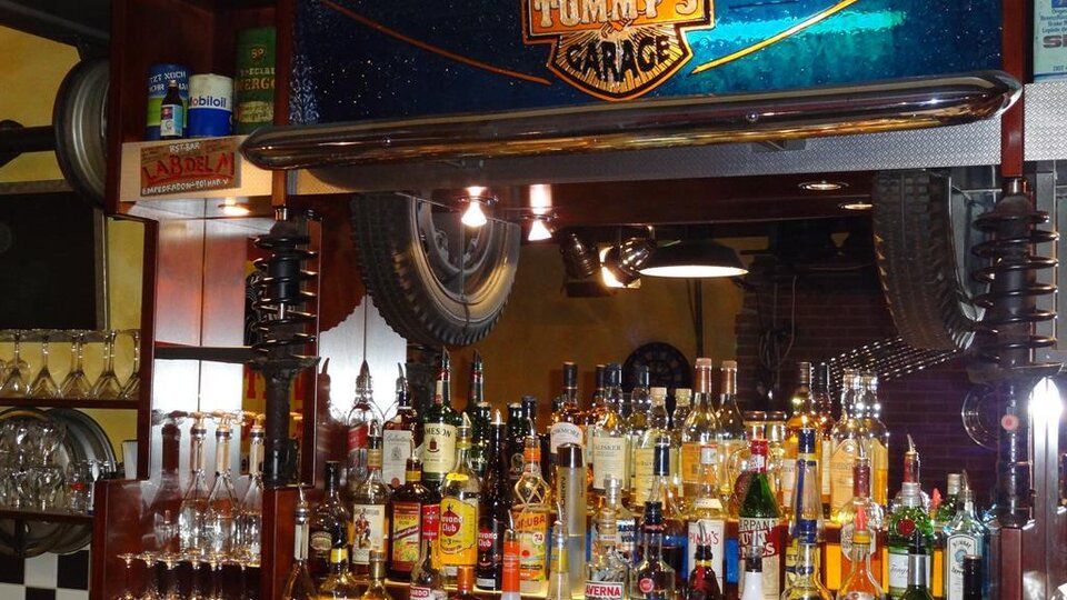 A bar named "Tommy's Garage," filled with various bottles of alcohol and glasses. Tires are hanging on the wall, and the lighting is cozy.