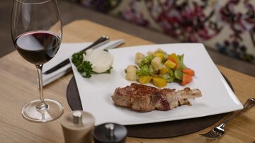 An elegant plate with a piece of meat, colorful vegetables, and a dollop of mashed potatoes. A glass of red wine on a wooden table.