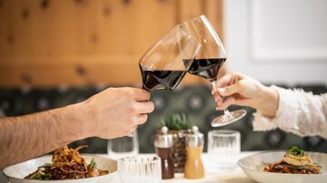 Two people are clinking wine glasses. In the background, there are plates with delicious dishes.