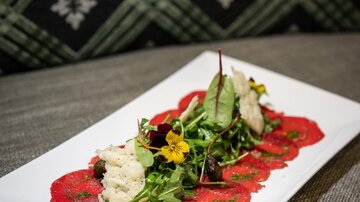 An elegant dish with red carpaccio, surrounded by fresh salad and edible flowers. The presentation is appealingly arranged on a white plate.