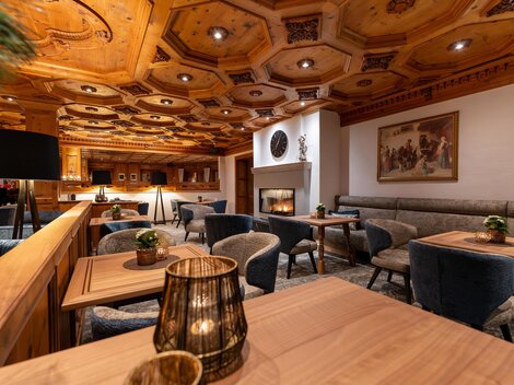 A cozy restaurant with wooden paneling and elegant furniture. There are tables, light-filled areas, and a fireplace in the background.