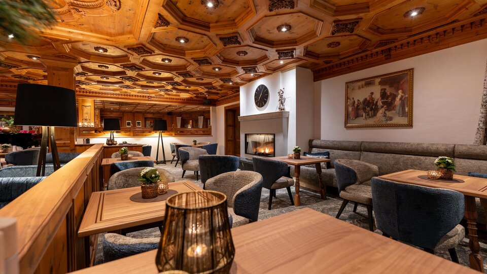 A cozy restaurant with wooden paneling and elegant furniture. There are tables, light-filled areas, and a fireplace in the background.