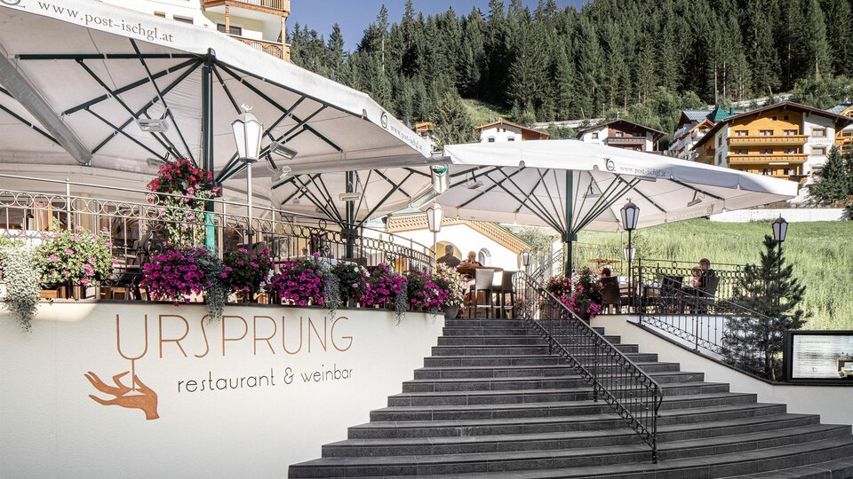 Ein gemütliches Restaurant mit Terrassen und blühenden Blumen. Im Hintergrund sind grüne Berge und traditionelle Hütten zu sehen.