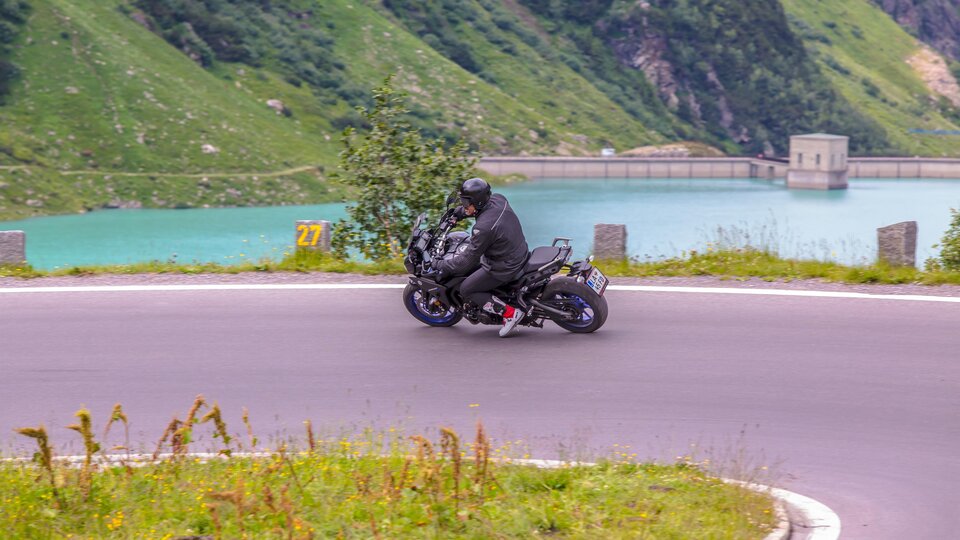 Ein Motorradfahrer fährt auf einer kurvenreichen Straße entlang eines klaren Sees. Die grüne Landschaft um ihn herum schafft eine beeindruckende Kulisse.