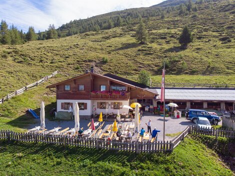 Ein alpenländliches Gasthaus mit Terrasse und Liegestühlen, umgeben von grünen Wiesen. Im Hintergrund erheben sich sanfte Hügel und Bäume.