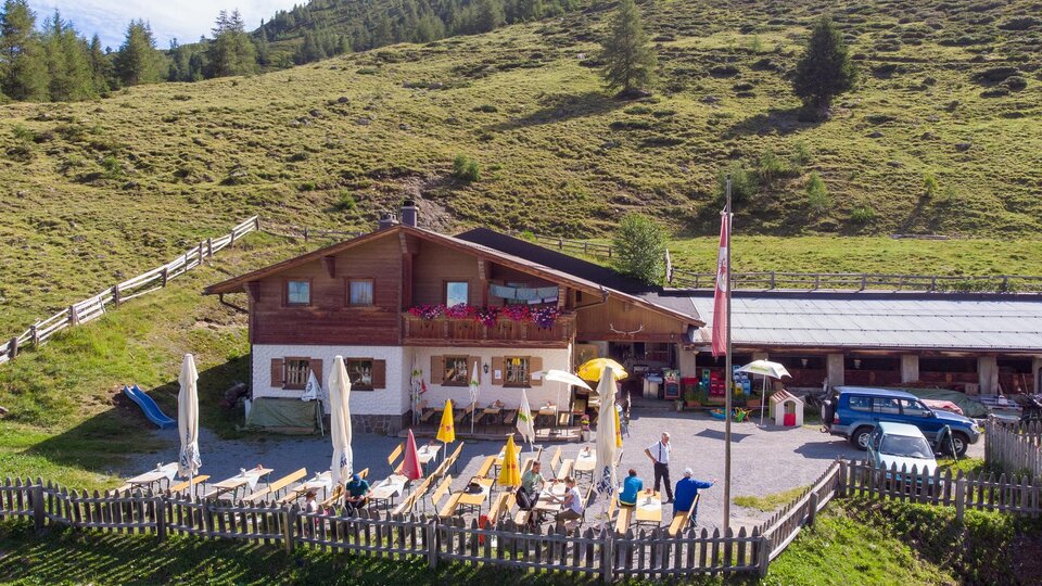 Ein alpenländliches Gasthaus mit Terrasse und Liegestühlen, umgeben von grünen Wiesen. Im Hintergrund erheben sich sanfte Hügel und Bäume.