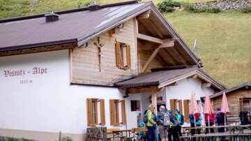 A cozy alpine lodge with a wooden facade and several outdoor tables. Hikers are standing in front of the entrance and enjoying the mountain landscape.
