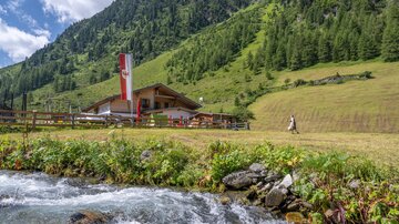 A picturesque alpine hut surrounded by green meadows and high mountains. A clear stream flows in the foreground.