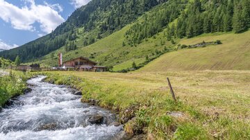 A clear stream flows through a green meadow, surrounded by gentle hills and wooded mountains. In the background, a rustic house is visible.