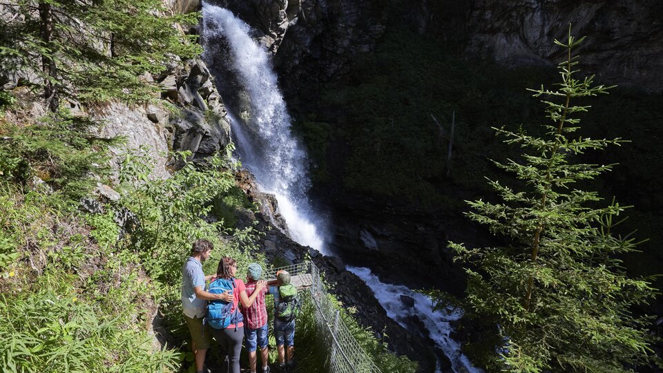 A group of people stands by a waterfall in nature. Surrounded by trees and lush vegetation, they enjoy the view.