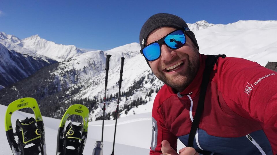 Ein Mann macht ein Selfie im Schneegebirge. Im Hintergrund sind verschneite Berge und Schneeschuhe zu sehen.