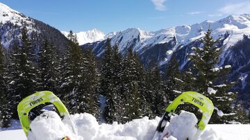 Schneeschuhe stehen in frischem Schnee, umgeben von einer malerischen Berglandschaft. Im Hintergrund sind hohe Berge und ein klarer blauer Himmel zu sehen.