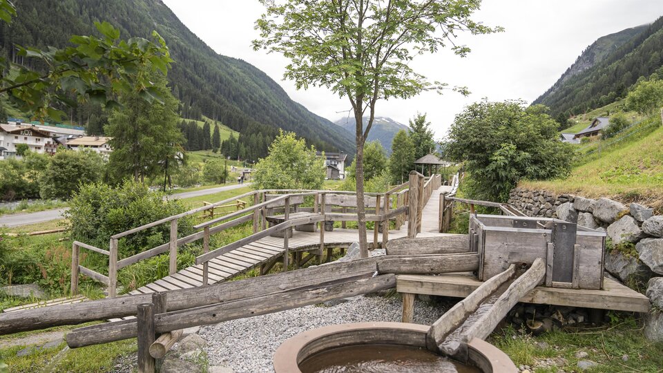 Eine ruhige Landschaft mit einem Holzsteg und einer Wasserstelle. Umgeben von grünen Hügeln und Bäumen.