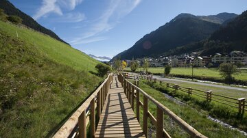Ein schöner Holzsteg führt durch eine grüne Landschaft mit Bergen im Hintergrund. Das Wetter ist klar und die Natur strahlt Ruhe aus.