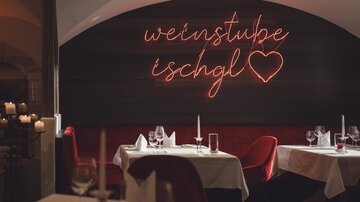 Ein stilvoller Restaurantbereich mit gedeckten Tischen, roten Samtsitzen und einem warm leuchtenden Neon-Schriftzug „Weinstube Ischgl“ vermittelt eine elegante, gemütliche Atmosphäre. | © Hannes Niederkofler