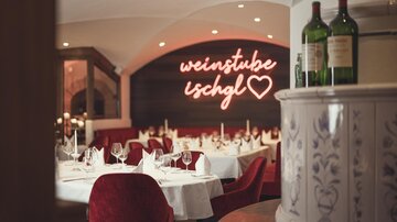 Ein eleganter Restaurantbereich mit weiß gedeckten Tischen, roten Samtsesseln und dem warm leuchtenden Neon-Schriftzug „Weinstube Ischgl“ schafft eine stilvolle, gemütliche Atmosphäre. | © Hannes Niederkofler