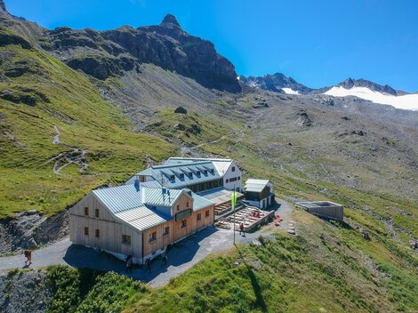 A mountain cabin surrounded by green meadows and mountains. The sky is clear and blue, perfect for an outdoor vacation.